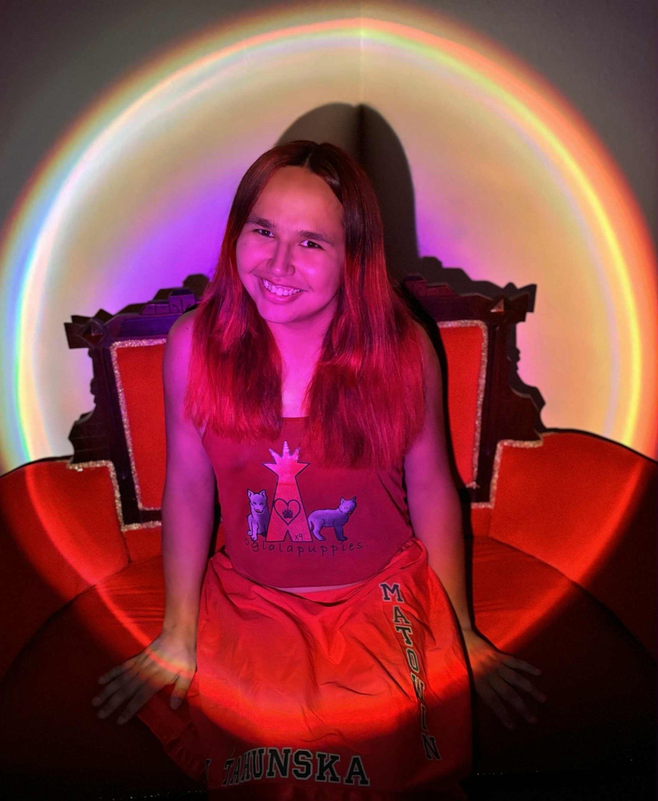 Nyna Matowin seated under a ring light, wearing an oglalapuppies shirt and a MATOWIN / TAHUNSKA wrap